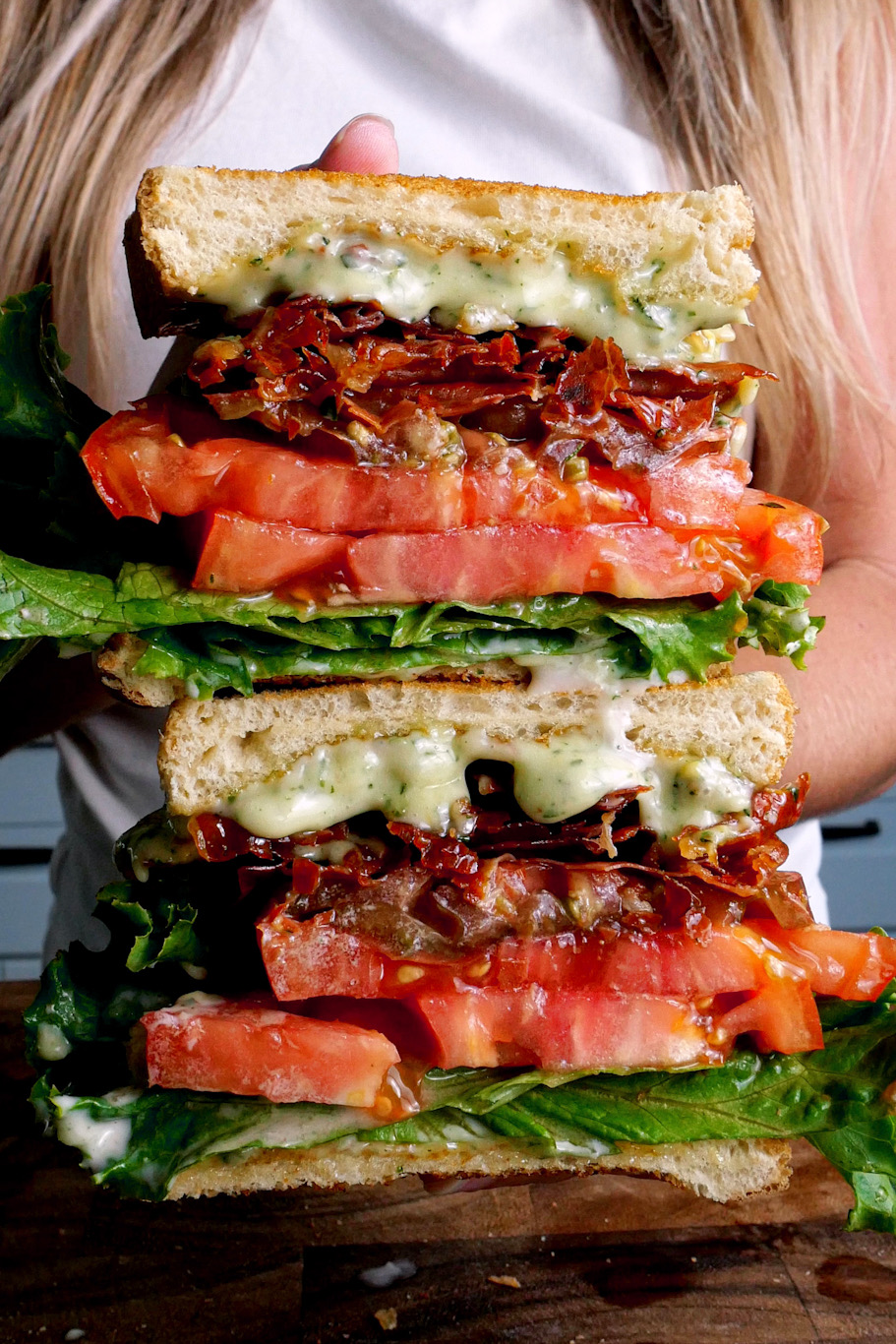 A cross-section of a "PLT" sandwich showcasing layers of crispy, toasted sandwich bread, vibrant green leaf lettuce, juicy heirloom tomato slices sprinkled with flaky salt, and savory prosciutto. The sandwich is generously spread with a creamy aioli made from mayo, collagen, lemon juice and zest, basil, tarragon, and minced garlic, adding a rich and tangy flavor to each bite.